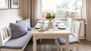 A dining area with a table set for four, a bench with cushions, and a window with a view of houses. - Haus Südstrand Whg. 174 - Haus Südstrand, Gemütliche 2-zimmerwohnung auf ca. 38 m² für bis zu 4 (Sylt)
