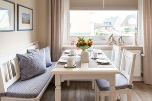 Un coin repas avec une table dressée pour quatre personnes, un banc agrémenté de coussins et une fenêtre donnant sur des maisons.