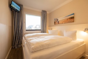 Une chambre d’hôtel avec un lit, un téléviseur fixé au mur, une fenêtre avec des rideaux et un tableau accroché au mur.