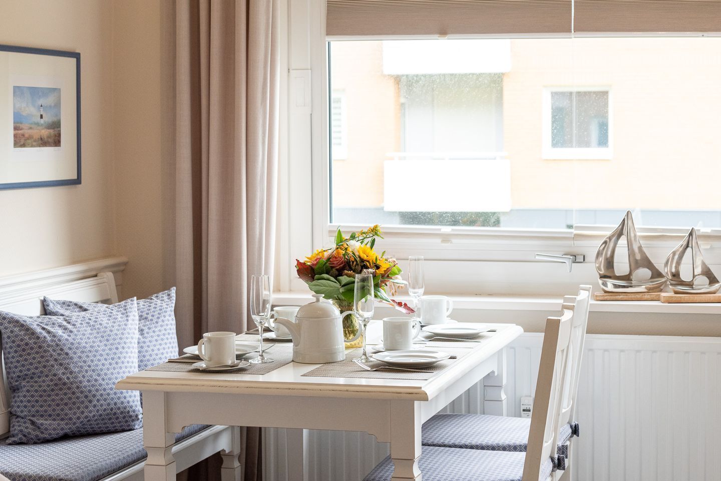 A dining area with a table set for two, a vase of flowers, and a view of the sea through the window.