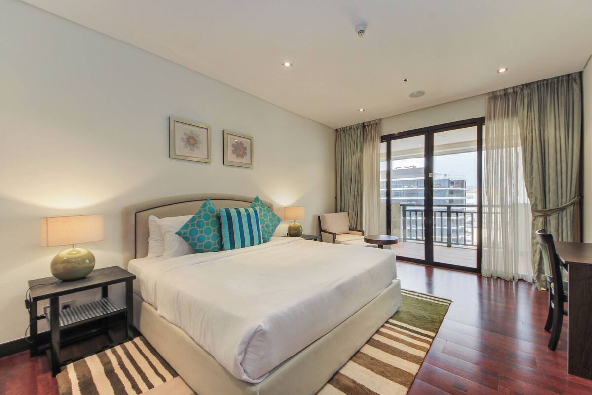 A hotel room with a large bed, a desk, a chair, a view of the city, and a balcony.