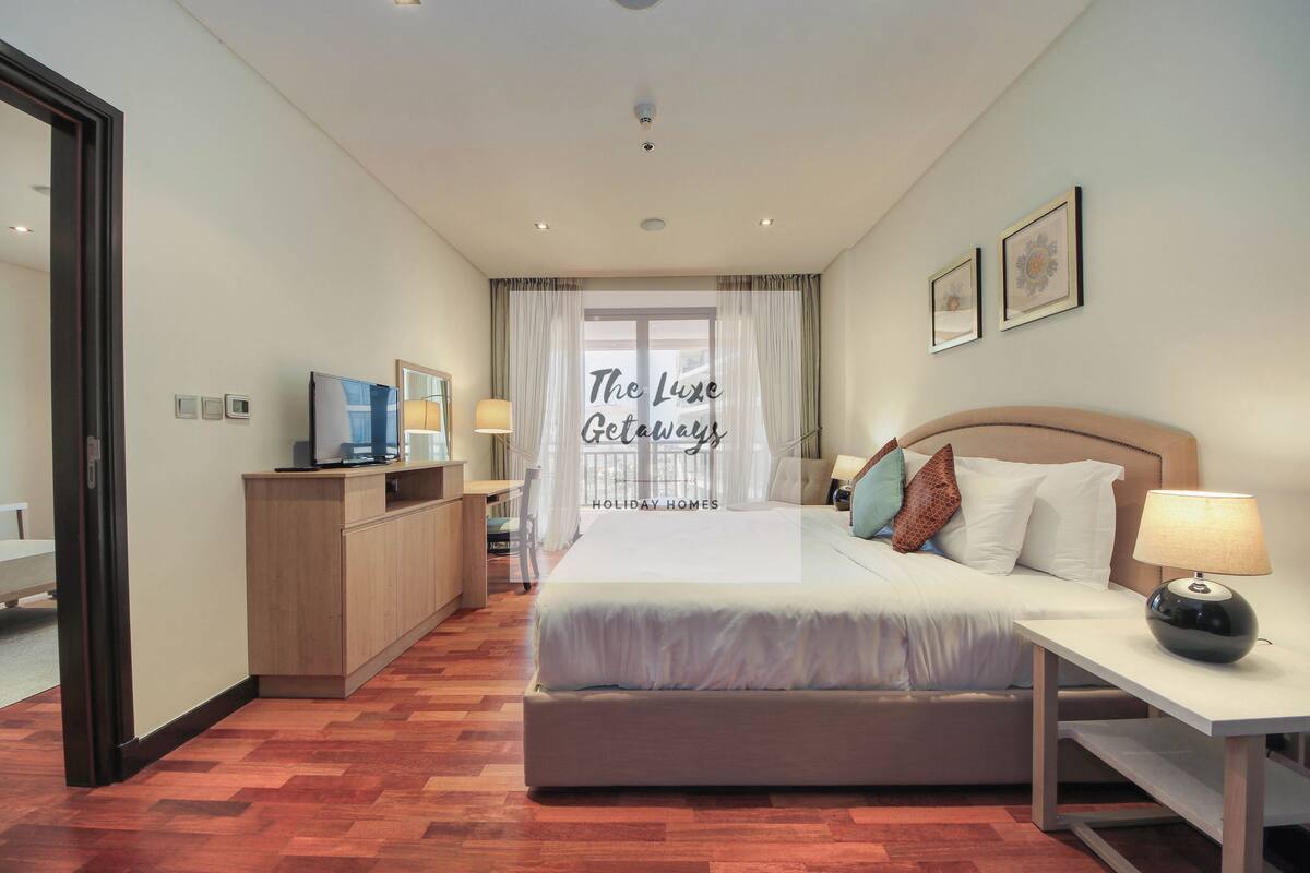 A hotel room with a bed, bedside table, lamp, and a desk with a computer.
