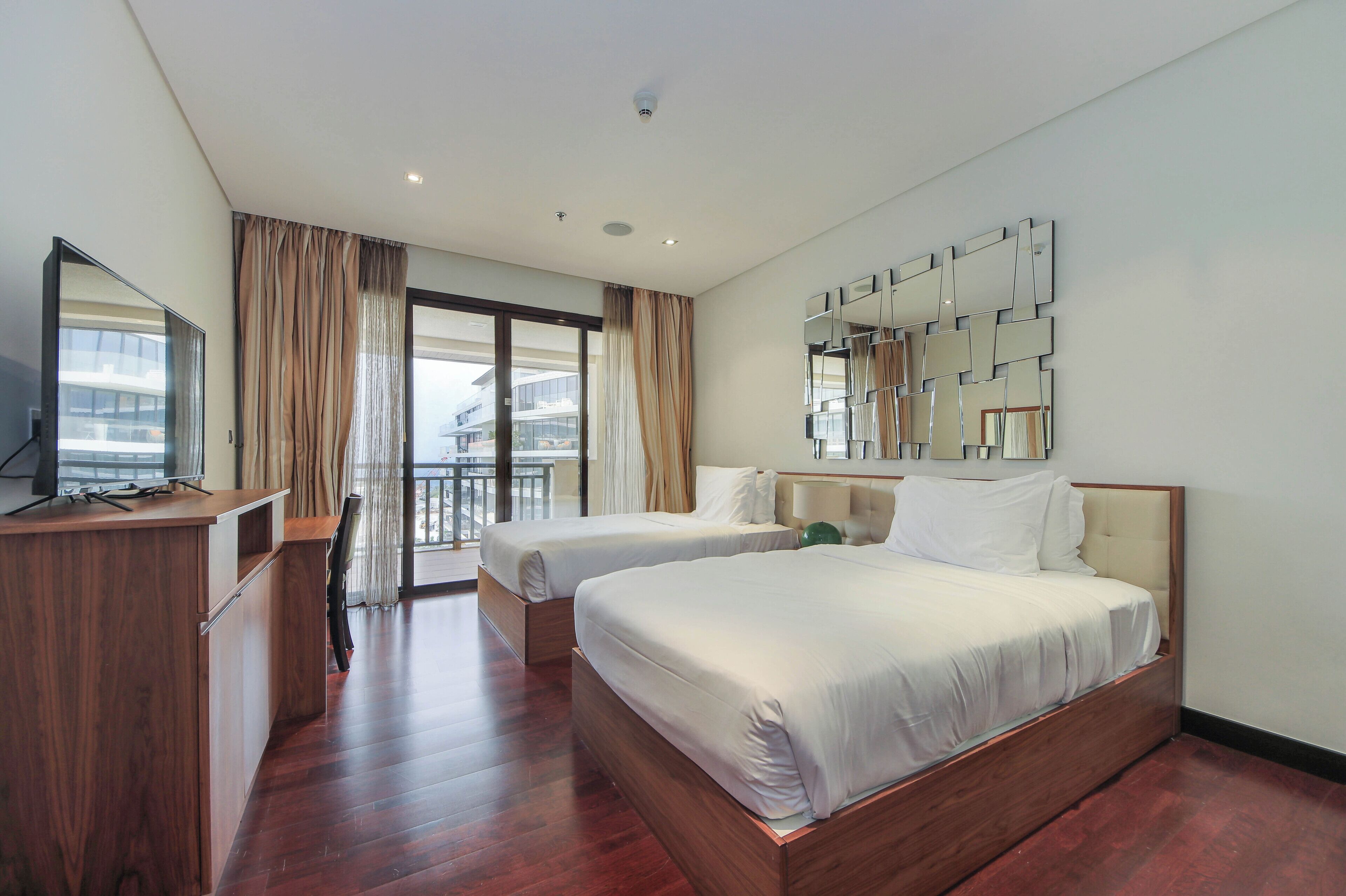 A hotel room with two beds, a wooden desk, a large mirror, and a balcony with curtains.