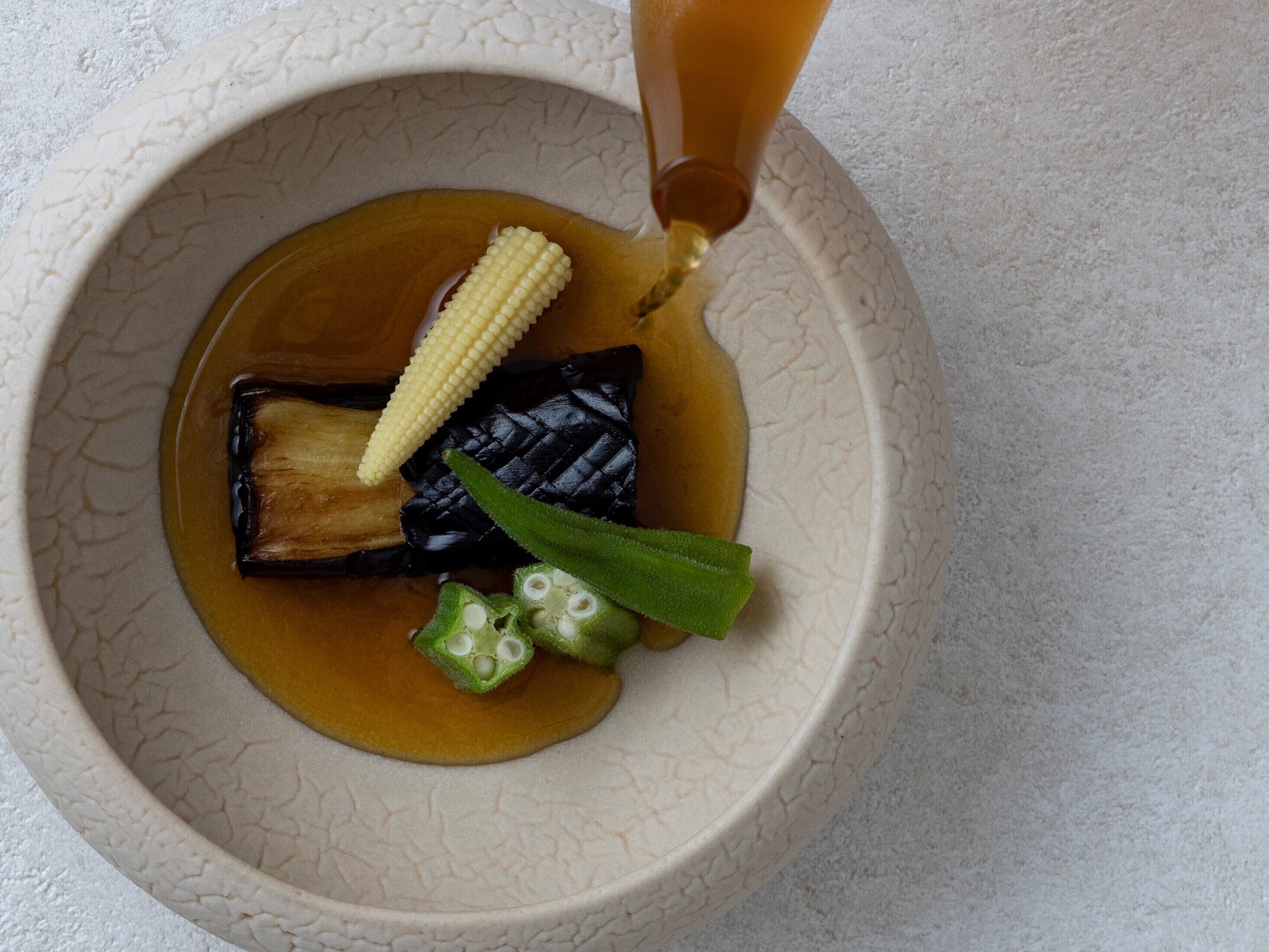 A bowl with a piece of grilled eggplant, a corn cob, and sliced okra in a clear liquid.
