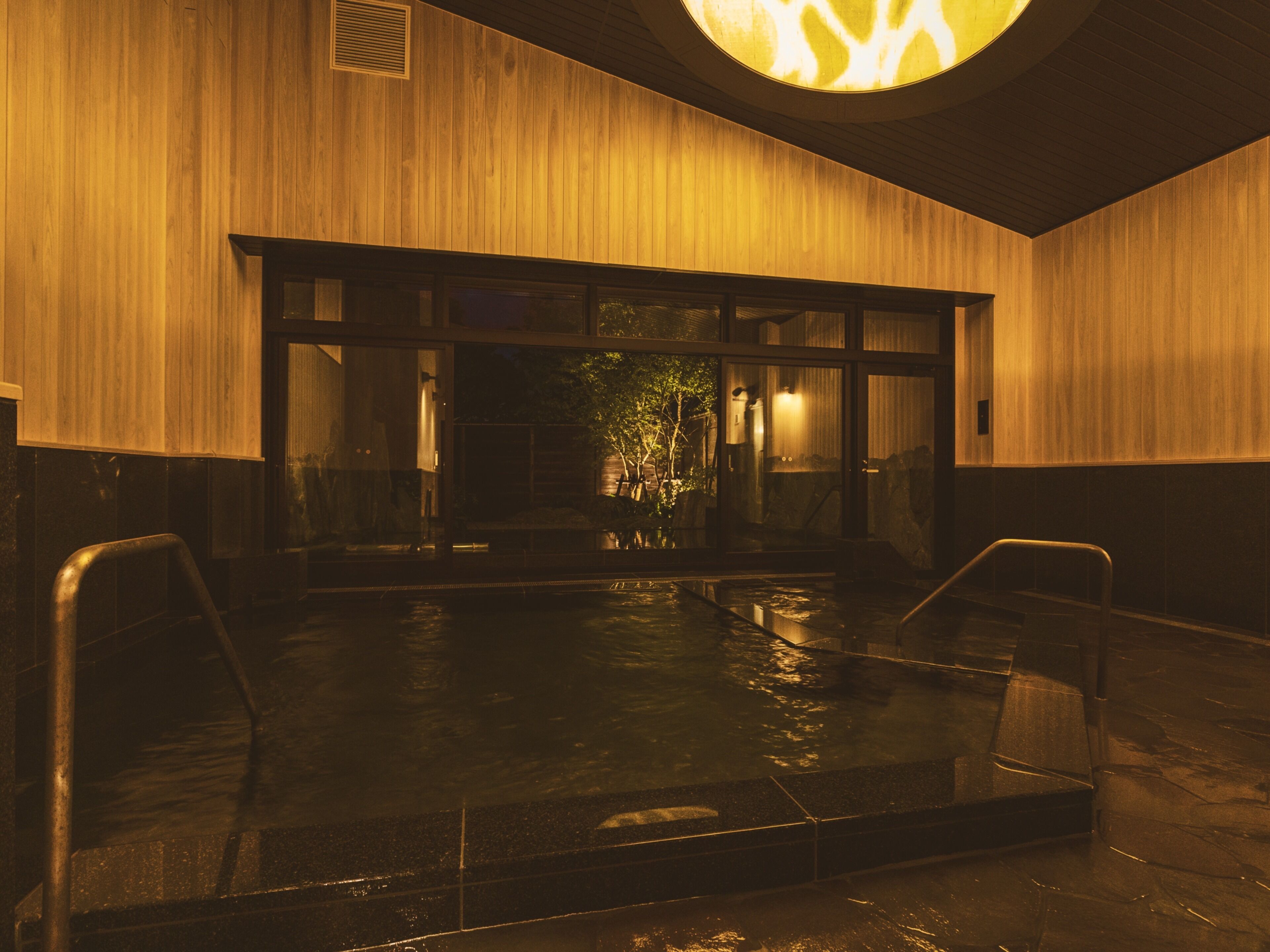 Un espace piscine intérieure chauffé, doté d’un mur en bois et d’une porte en verre donnant sur un jardin.