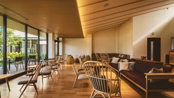 A spacious indoor area with wooden flooring, a long row of chairs, a sofa, and a large window providing a view of greenery outside.