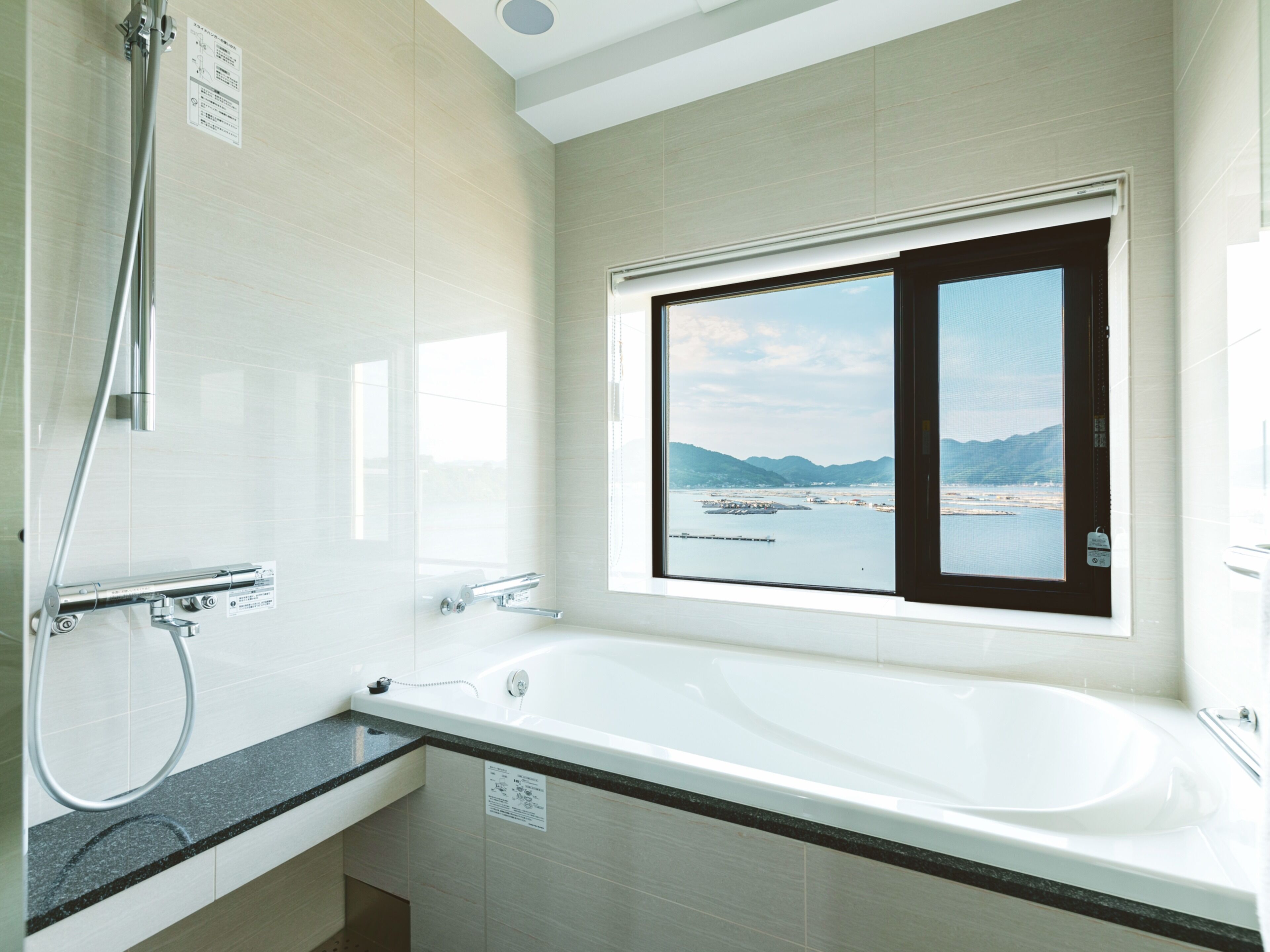 A modern bathroom with a large bathtub, a shower, and a window offering a view of a body of water and mountains.
