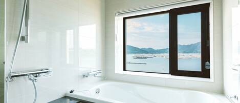 A modern bathroom with a large bathtub, a shower, and a window offering a view of a body of water and mountains.