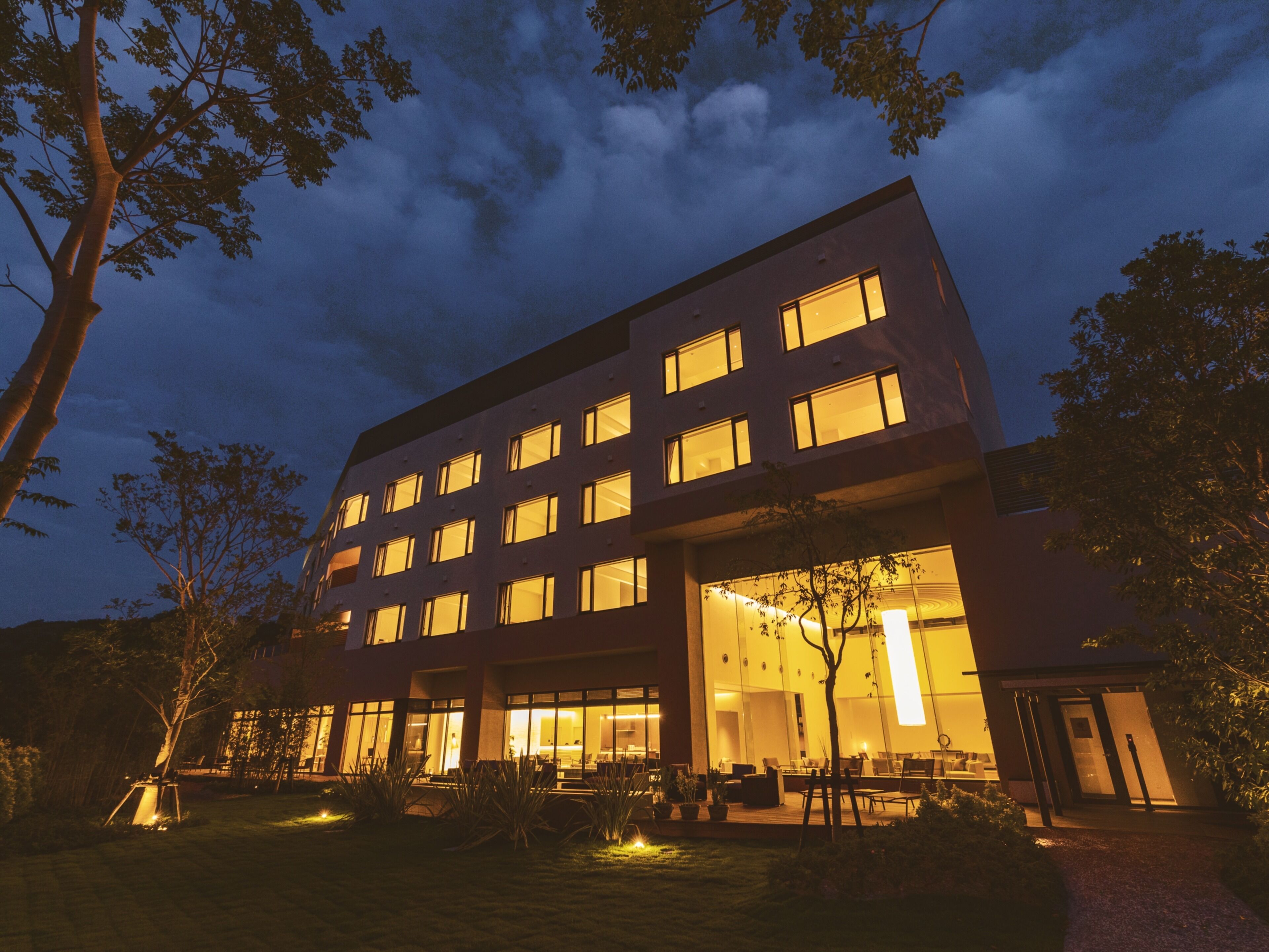 A modern building with multiple windows, a well-lit entrance, and a garden area with plants and seating.