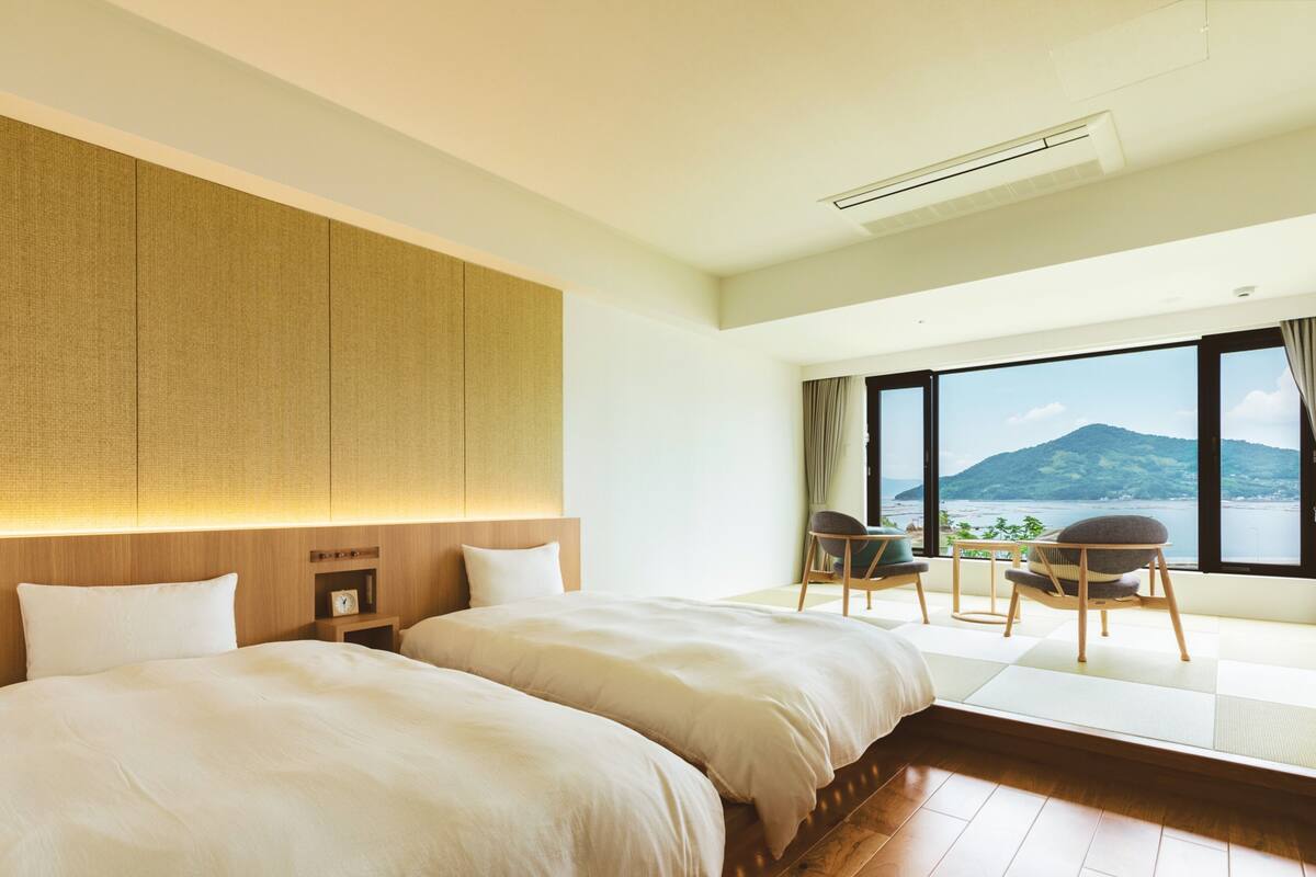 A hotel room with two beds, a wooden headboard, a nightstand, a chair, a view of a mountain and sea through large windows.