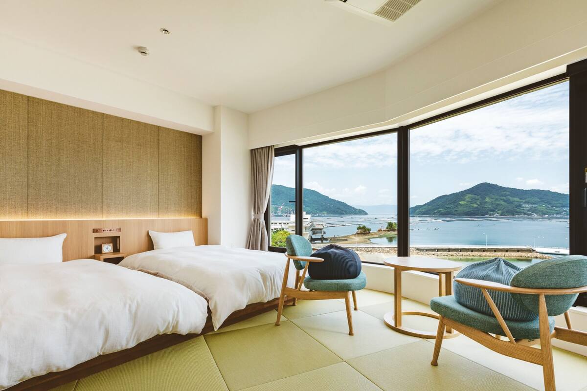 A modern hotel room with a large bed, two chairs, a small table, and a view of the sea and mountains.