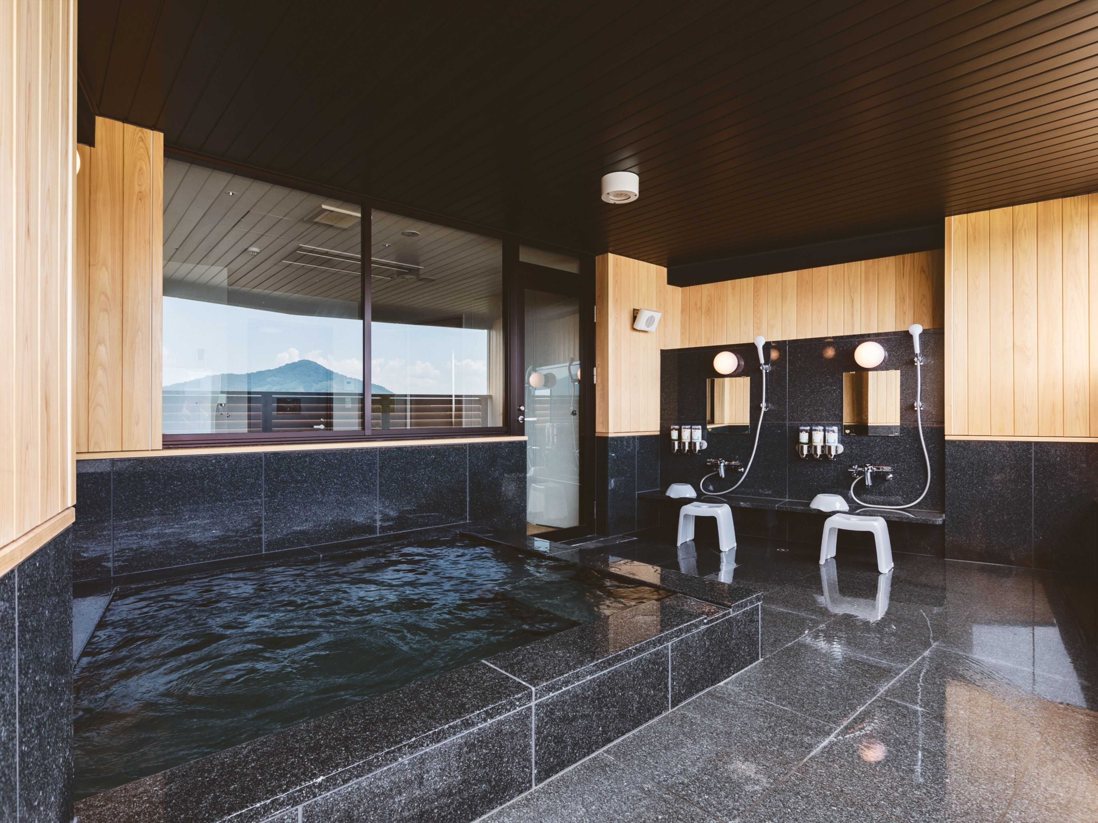 A modern bathroom with a large black marble sink, two white stools, wooden paneling, and a view of a mountain through a large window.