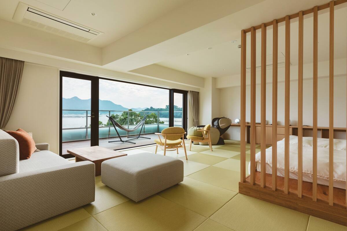 A modern hotel room with a large balcony, a sofa, a chair, a small table, and a bed.