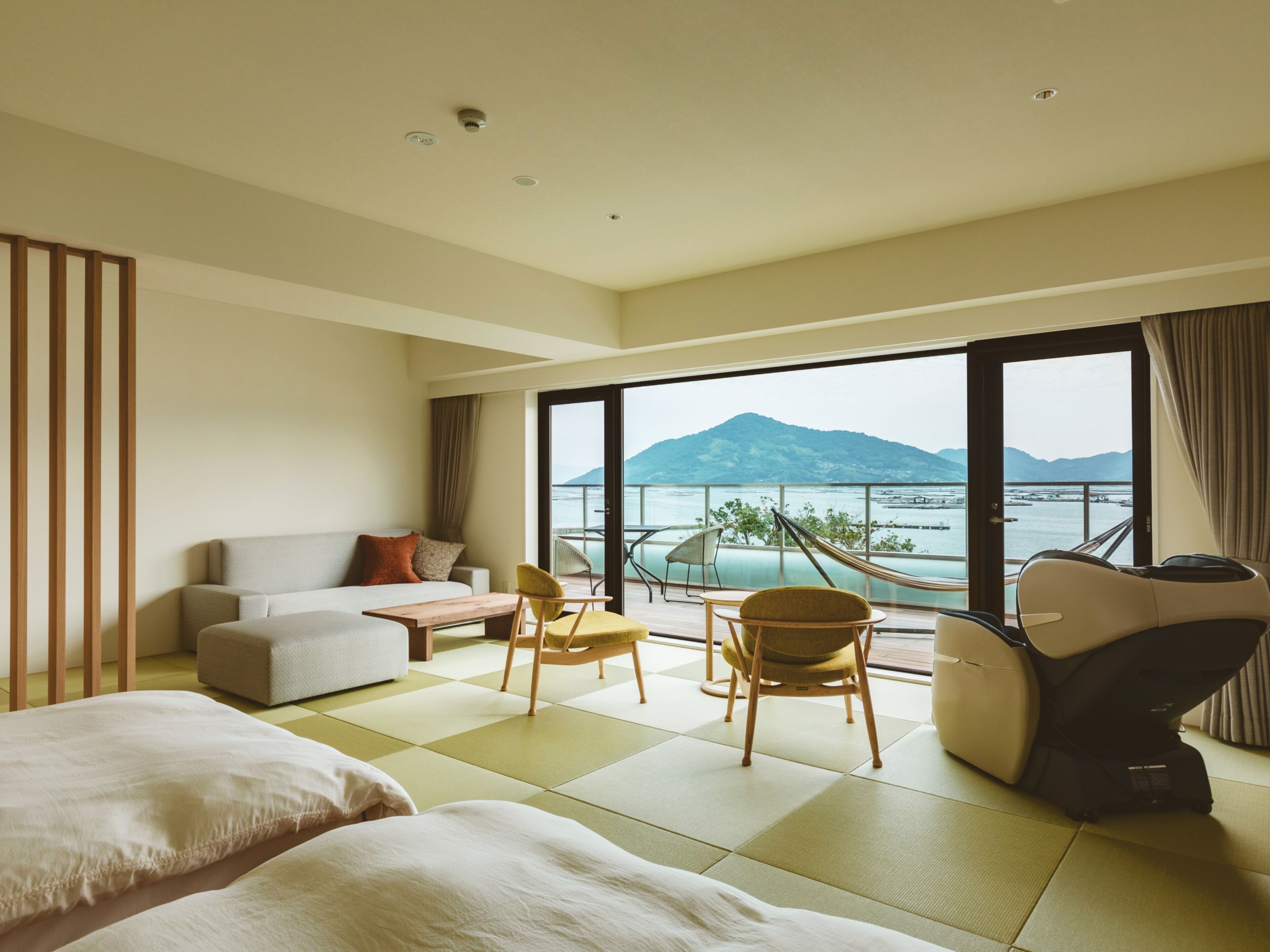 Une chambre d’hôtel moderne, dotée d’un canapé, de deux fauteuils, d’une petite table et offrant une vue sur la mer et les montagnes grâce à de grandes fenêtres.