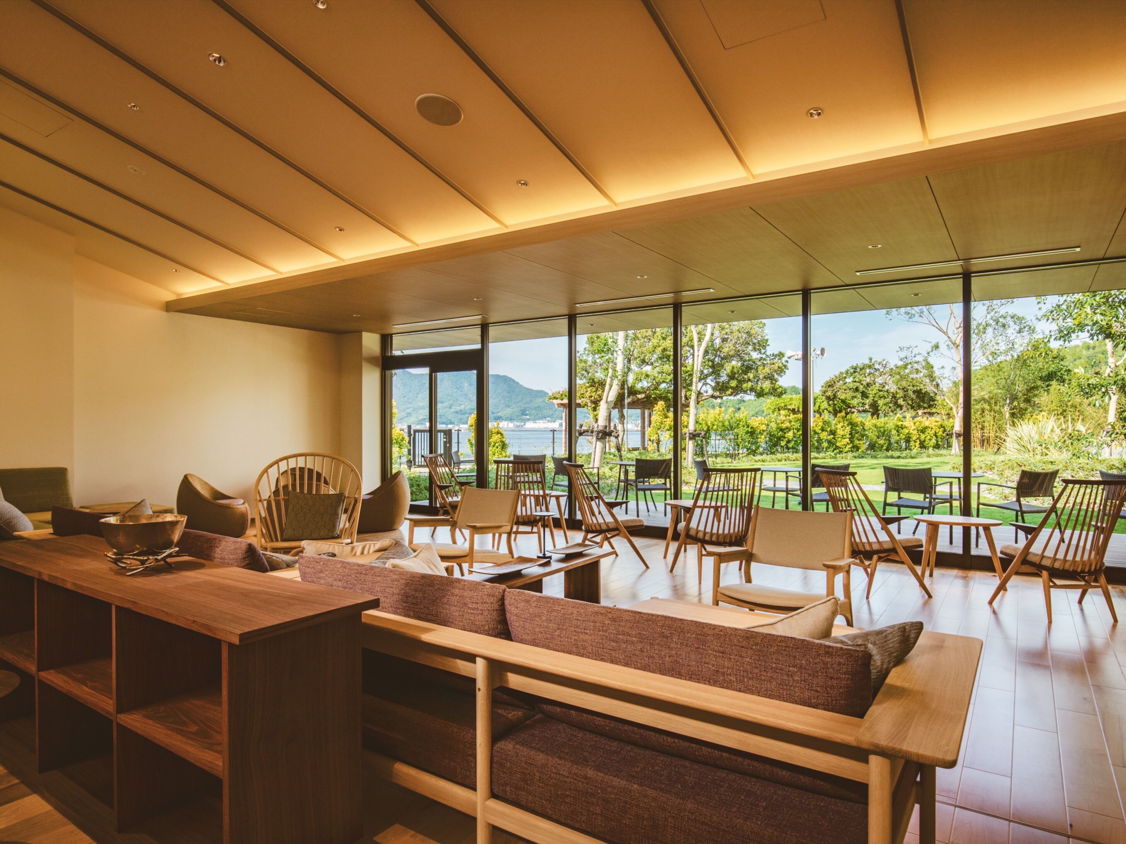 A modern interior with a wooden dining area, cushioned seating, and large windows offering a view of a lush garden and mountains.