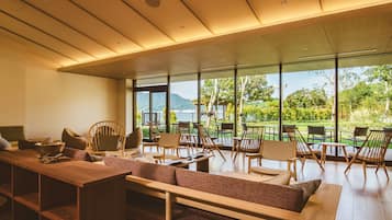 A modern interior with a wooden dining area, cushioned seating, and large windows offering a view of a lush garden and mountains.