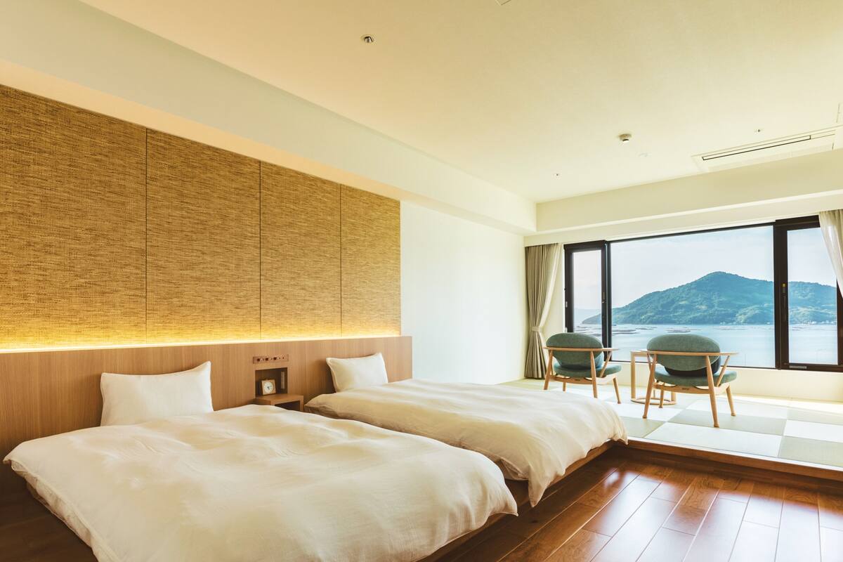 A hotel room with two beds, a wooden headboard, a small table with chairs, and a large window offering a view of a mountain and water.