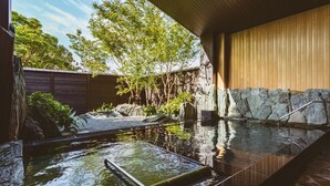 A spa area with a hot tub, stone walls, wooden paneling, and a view of trees. - Etajimasou (Etajima)