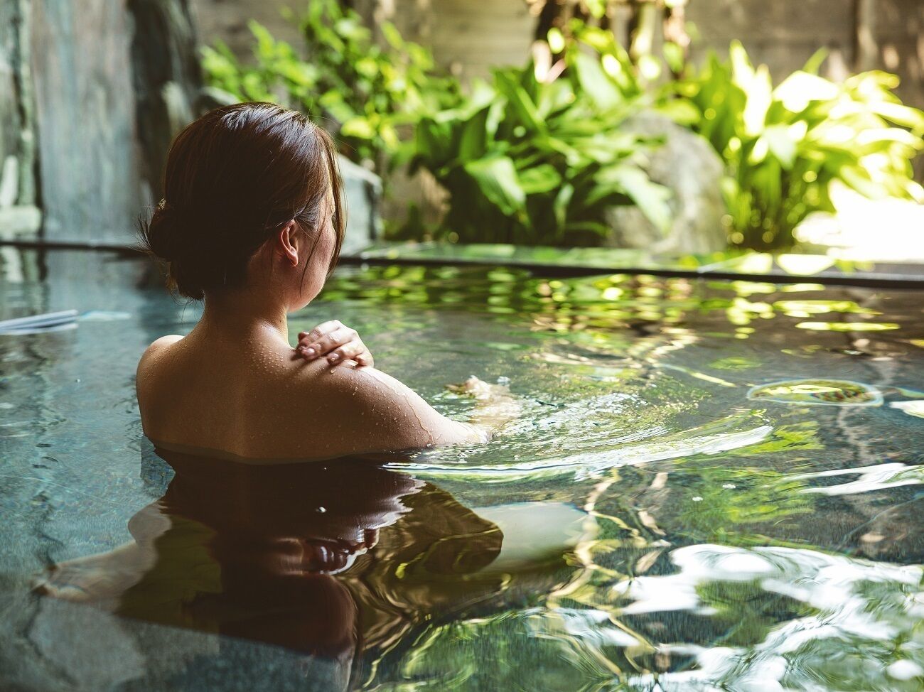 Una persona en una piscina interior transparente, con reflejos del agua y follaje al fondo.