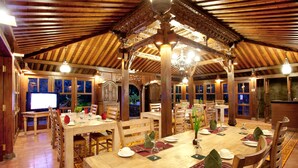 Un espace repas avec des tables et des chaises en bois, un lustre et une vue sur l’extérieur grâce à de grandes fenêtres.