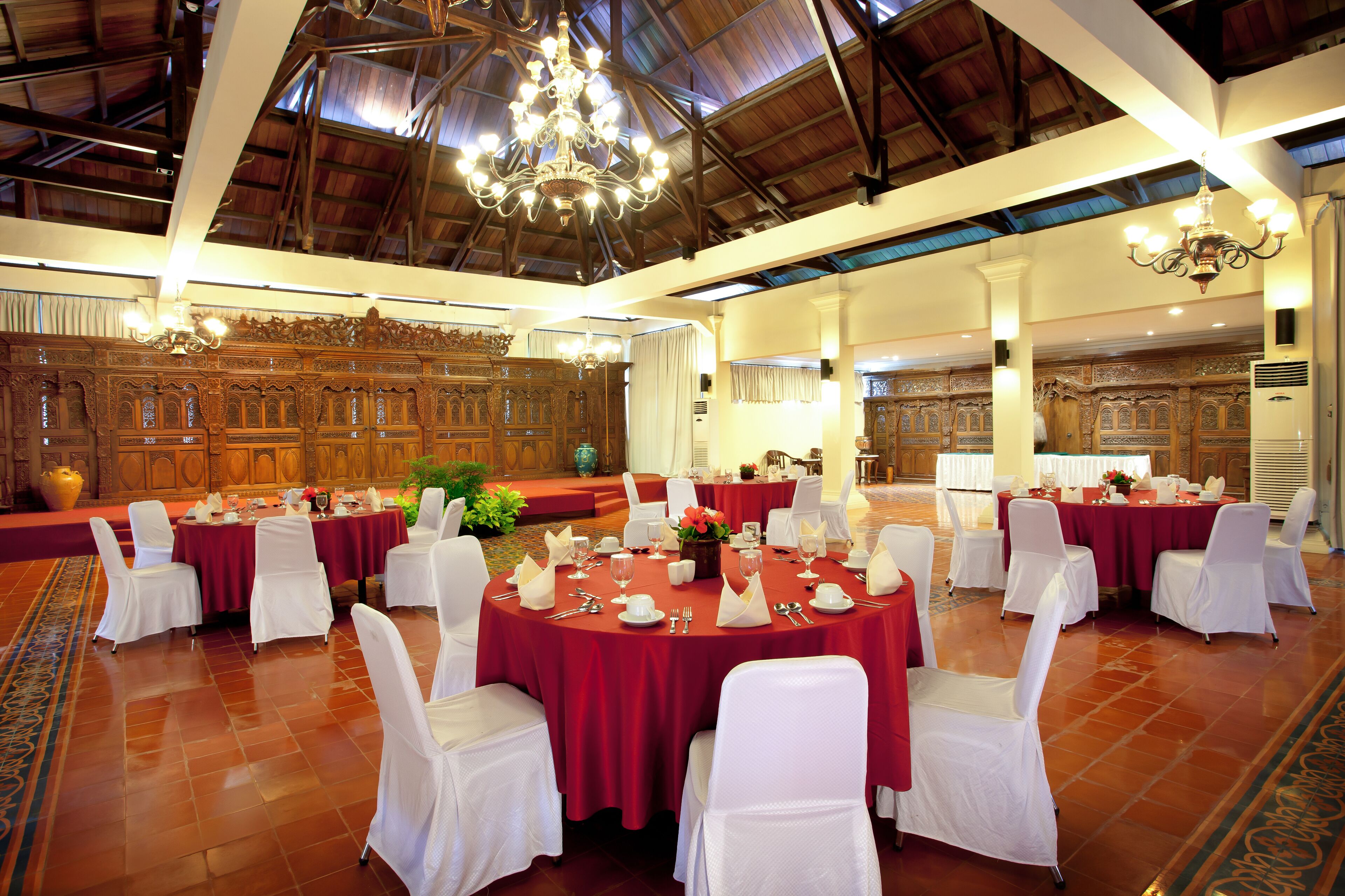 A banquet hall with round tables covered in red tablecloths, white chairs, and chandeliers.