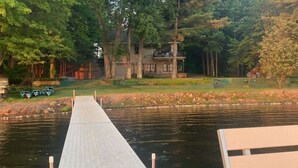 A wooden dock leading to a house by a river, surrounded by trees and a grassy area.