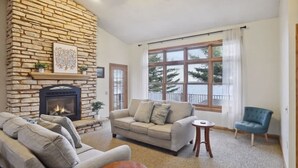 A living room with a stone fireplace, a beige sofa, a blue armchair, and a large window with a view of trees.