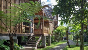 A wooden building with a staircase leading to a balcony, surrounded by lush greenery and a cobblestone path. - RUMAH KAYU RESORT (Ubud)