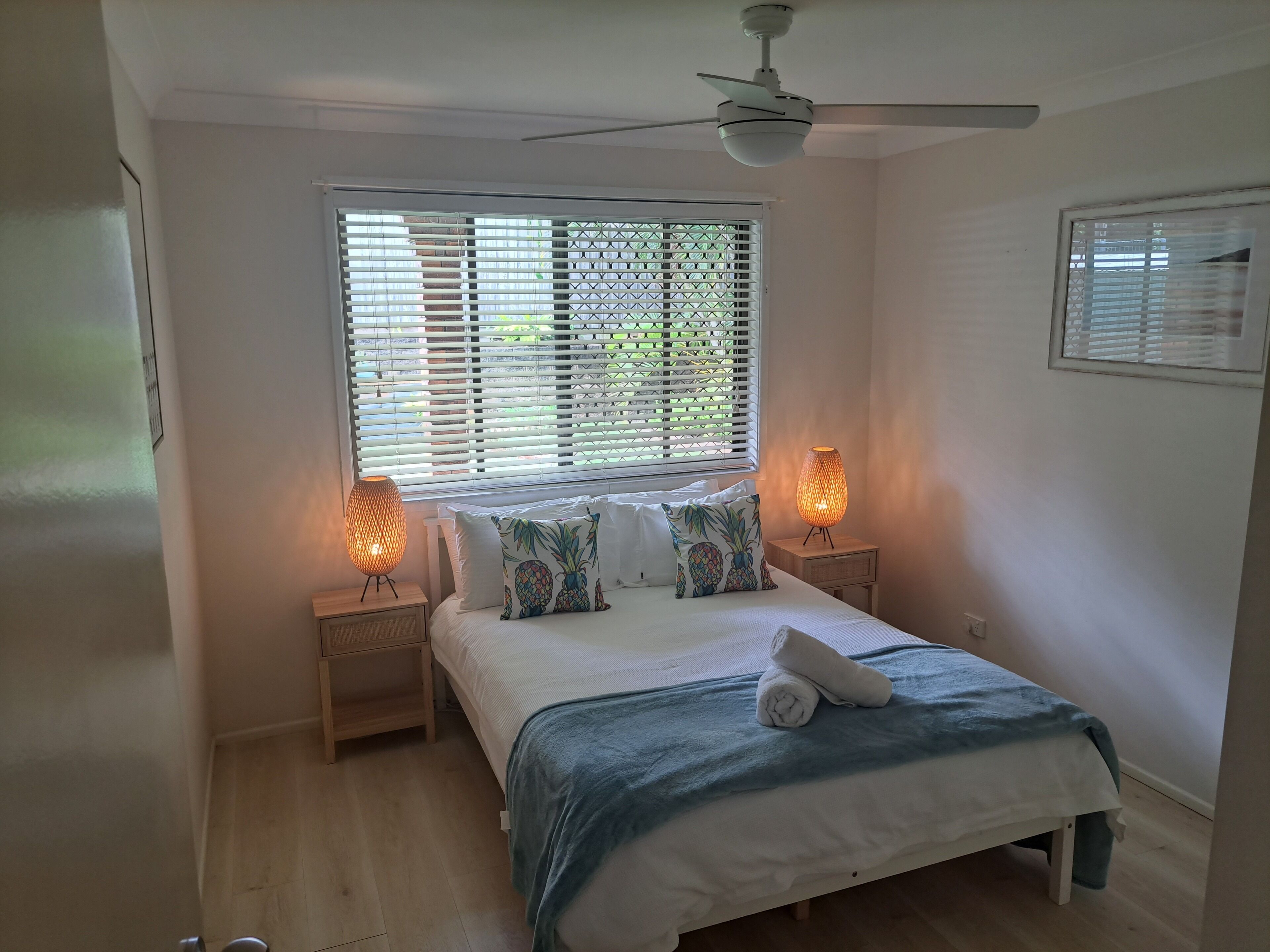 A bedroom with a bed, two bedside lamps, a ceiling fan, and a window with blinds.