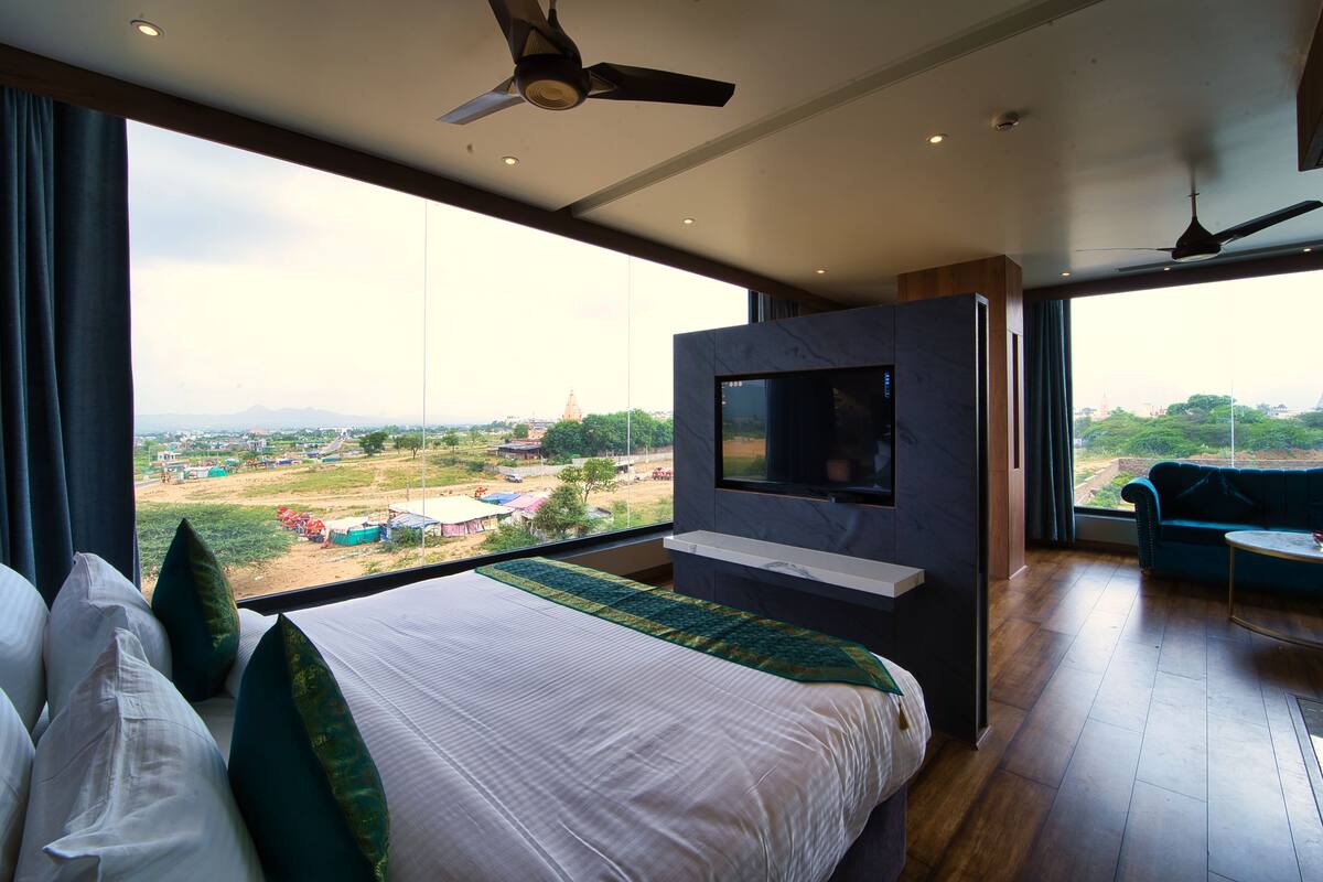 A modern hotel room with a large bed, a flat-screen TV mounted on the wall, and a balcony with a view of the countryside.