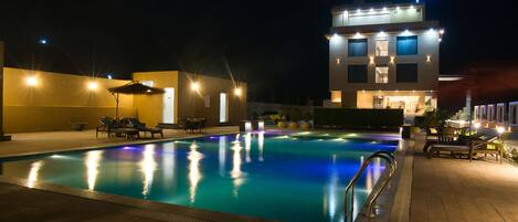 A swimming pool with a sunken seating area, surrounded by lounge chairs and umbrellas, with a building in the background.