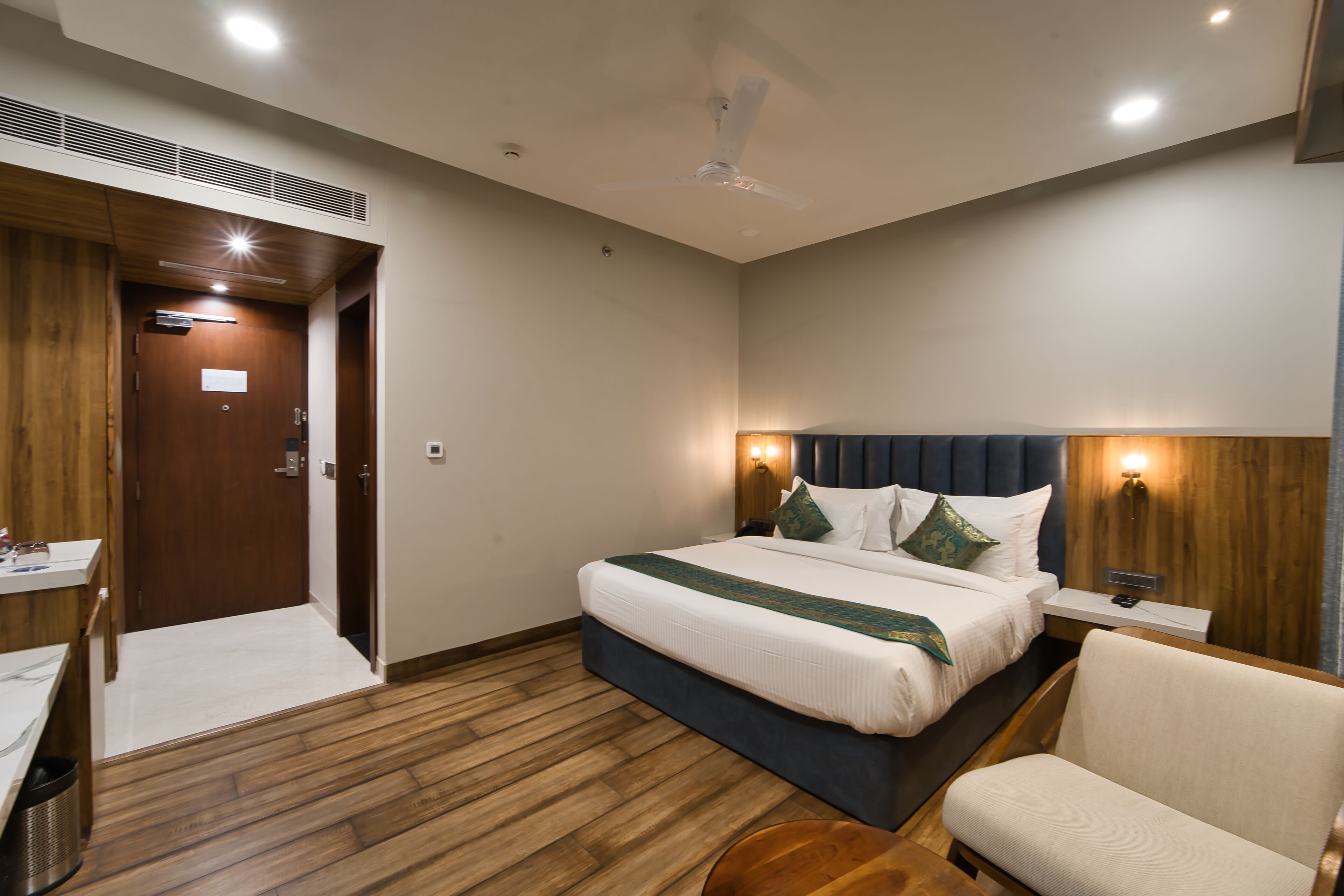 Habitación de hotel con piso de madera, cama con cabecera oscura, mesita de noche y una silla.