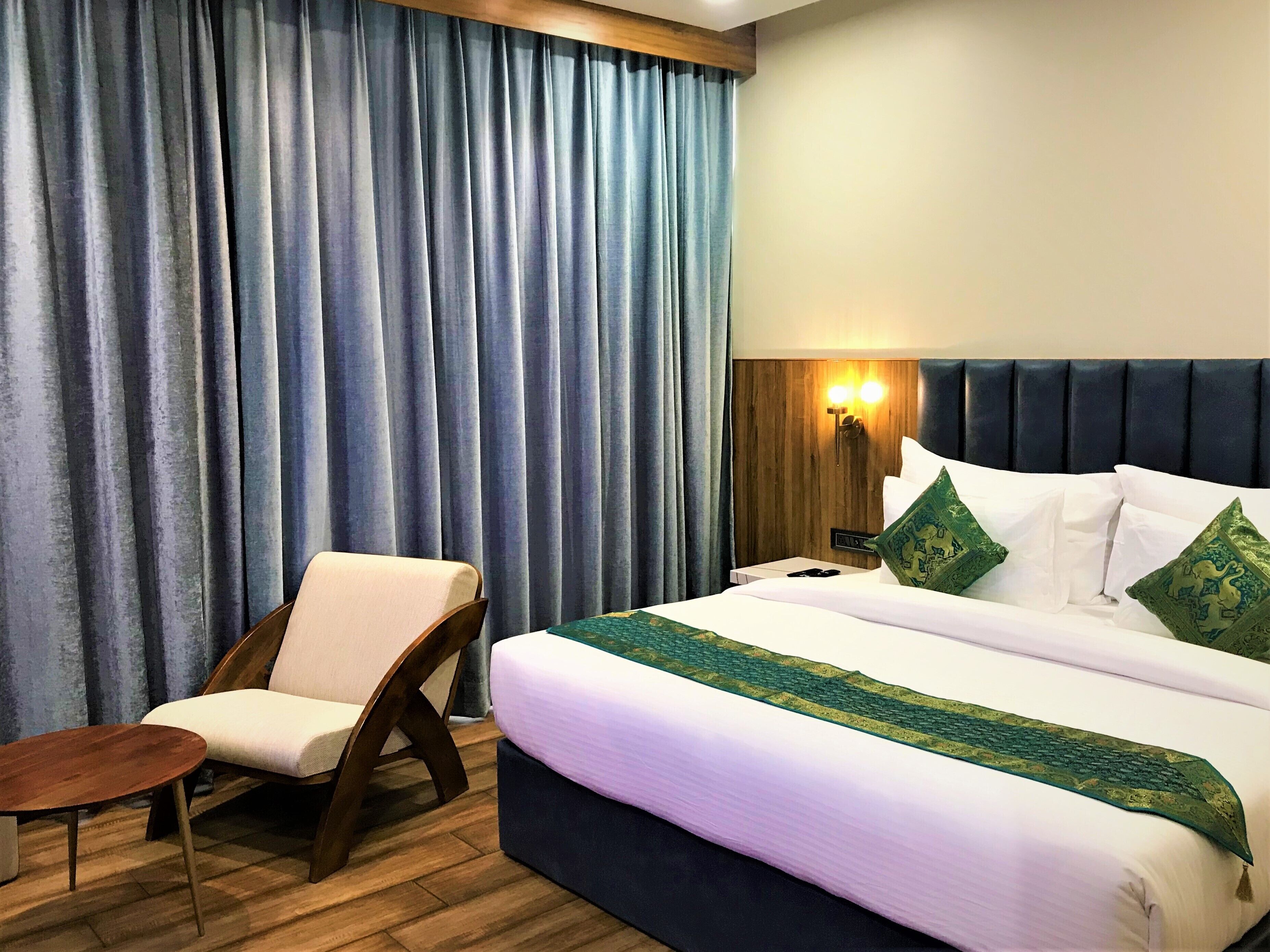 Habitación de hotel con cama, una silla, una mesita y un ventanal con cortinas.