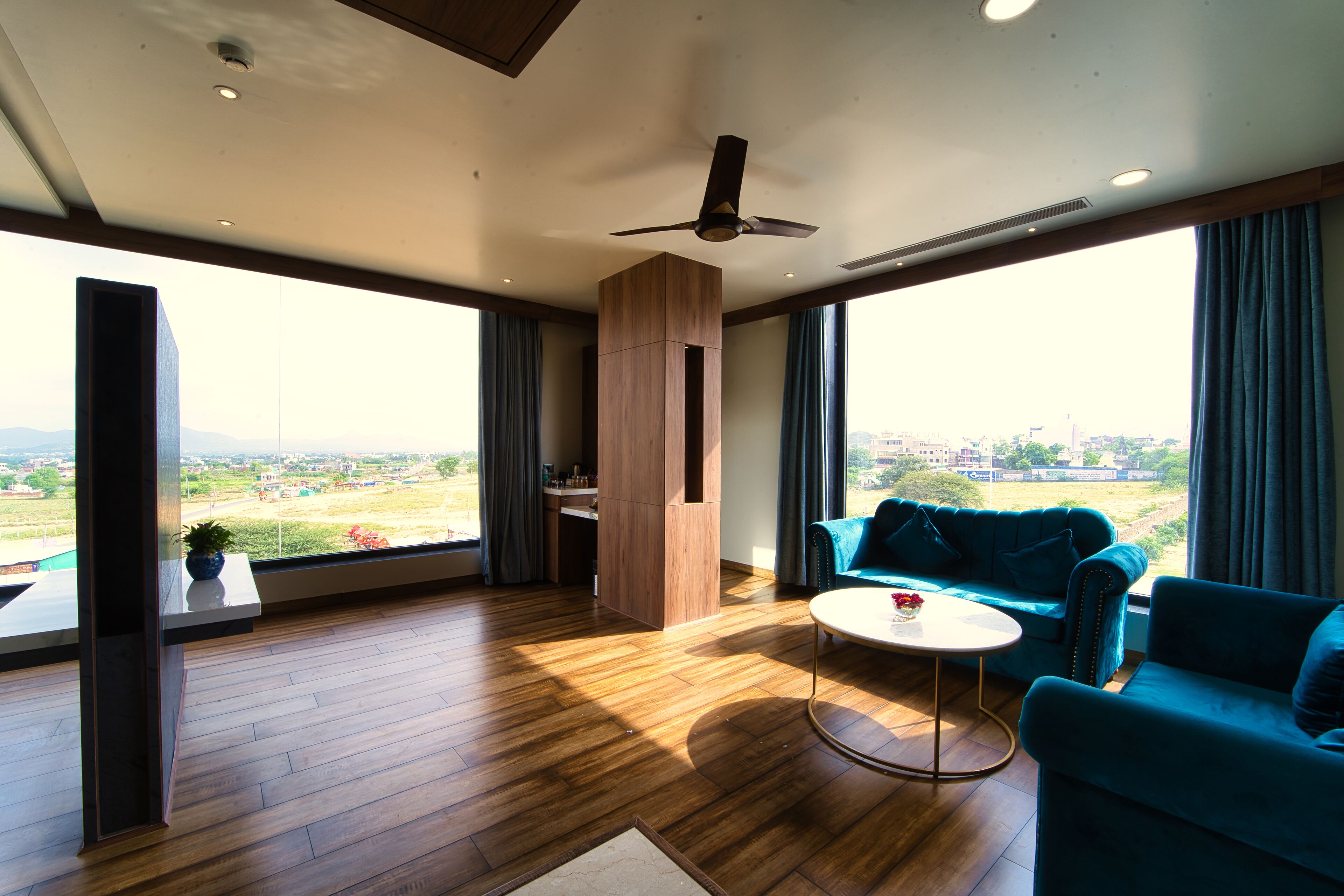 A spacious living area with a wooden floor, a ceiling fan, a blue sofa, a round coffee table, and large windows offering a view of a green landscape.