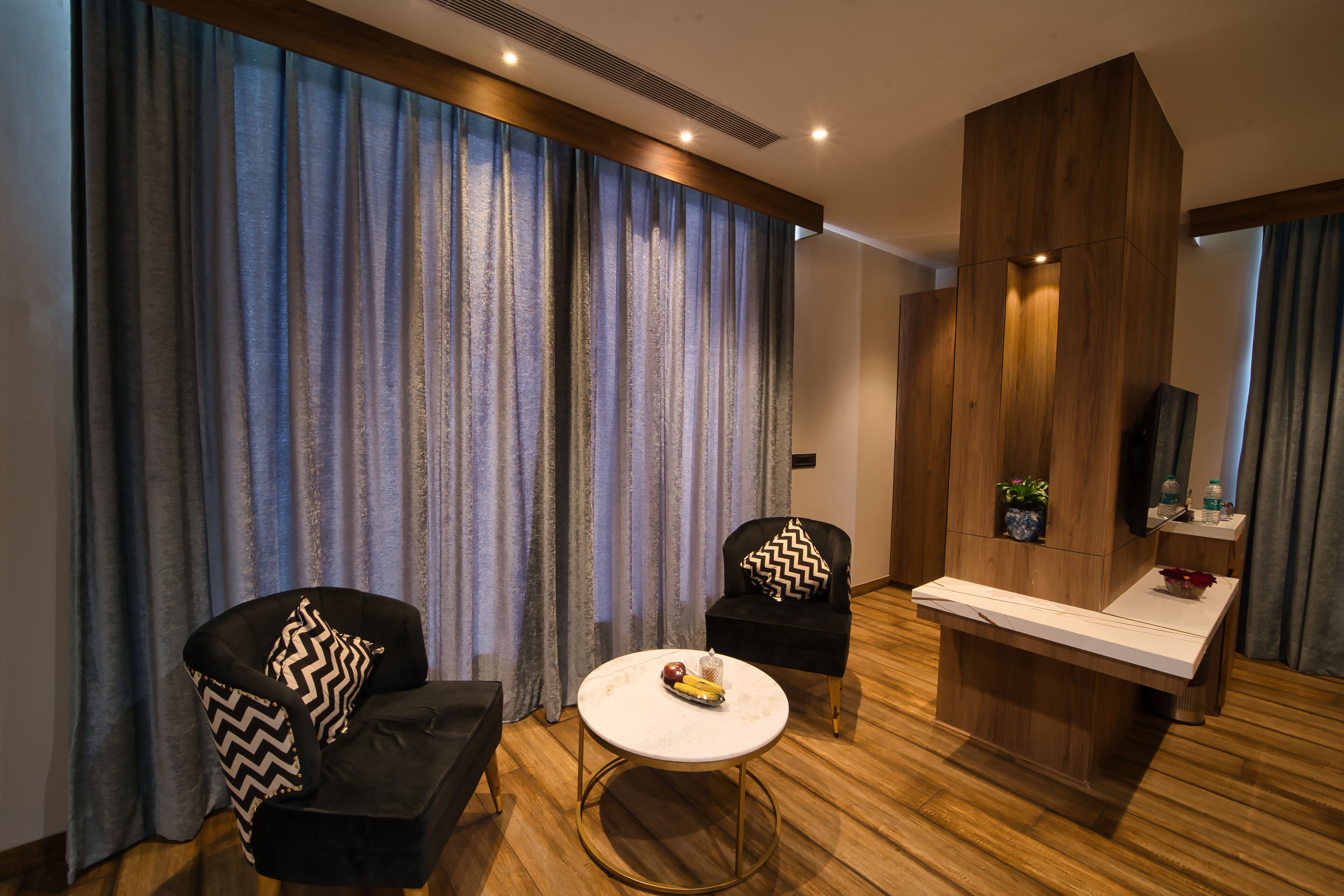 A modern hotel room with a wooden floor, two black armchairs with patterned cushions, a round white coffee table, and a wooden reception desk.