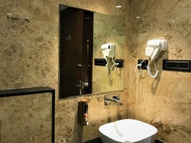 Un baño moderno con pared de mármol, un espejo grande, un lavamanos blanco y dos secadores de pelo fijados a la pared.