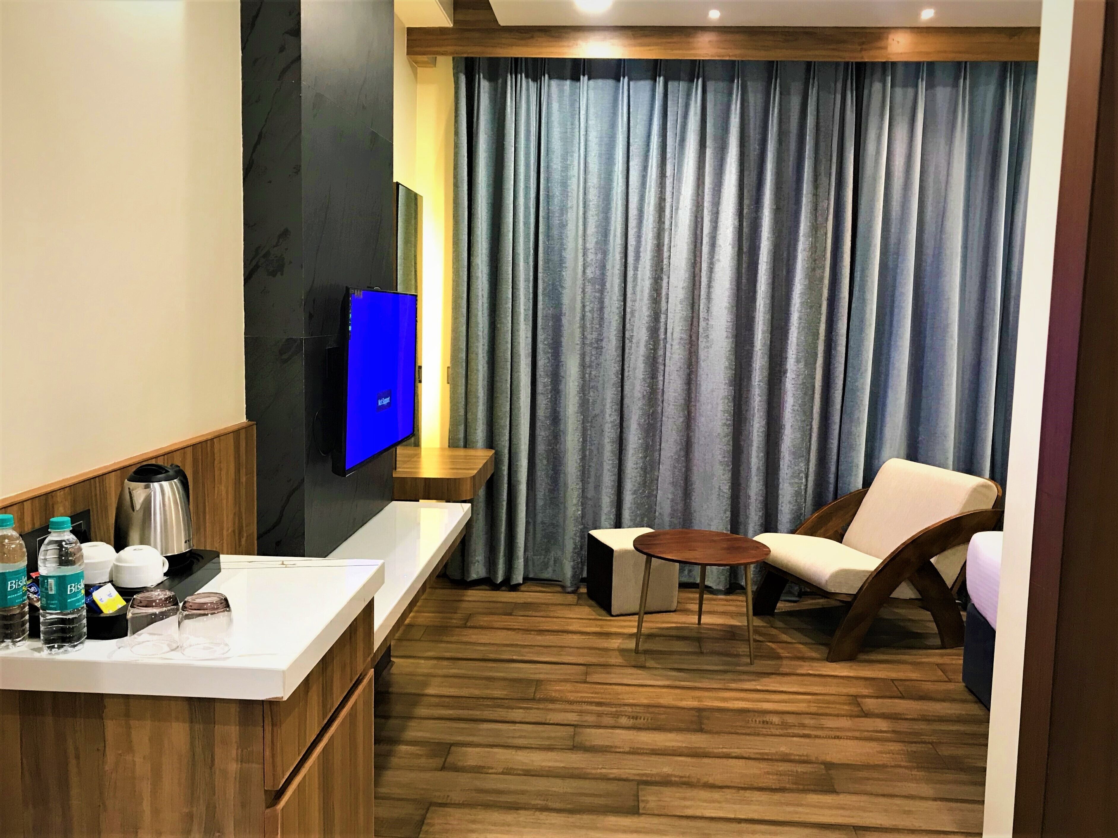 Ein Hotelzimmer mit Holzboden, einer weißen Ablagefläche mit Pflegeprodukten, einem Fernseher, einem Sessel und einem großen Fenster mit Vorhängen.