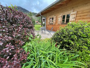 A wooden cabin with a patio area, surrounded by lush greenery and a mountain backdrop.