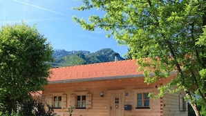 En timmerstuga med ett rött tegeltak, en veranda och en bergskedja i bakgrunden.