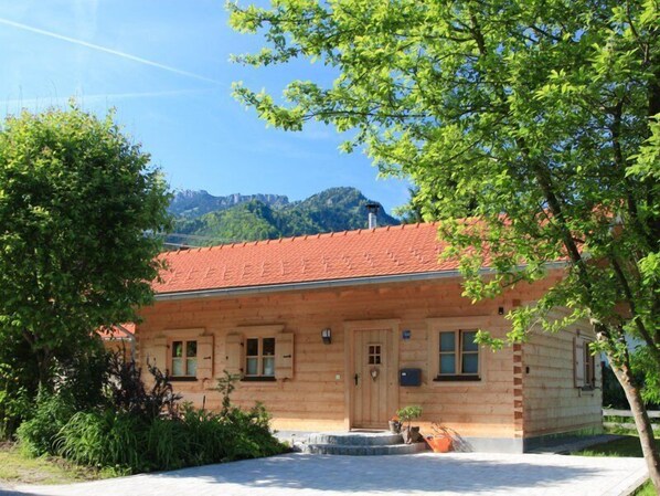 A wooden log cabin with a red-tiled roof, a front porch, and a mountain backdrop.