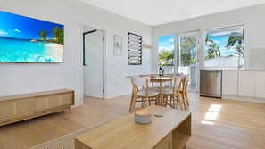Relax in a stylish living space with a large flat-screen TV, dining table, and sliding glass door leading to an outdoor area with a palm tree view.