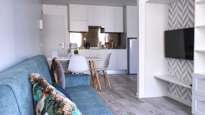 A modern living room with a blue sofa, a dining area with a white table and chairs, a kitchen with white cabinets, and a mounted TV. - Palacete da Baixa, Standard Studio Apartment (Faro)