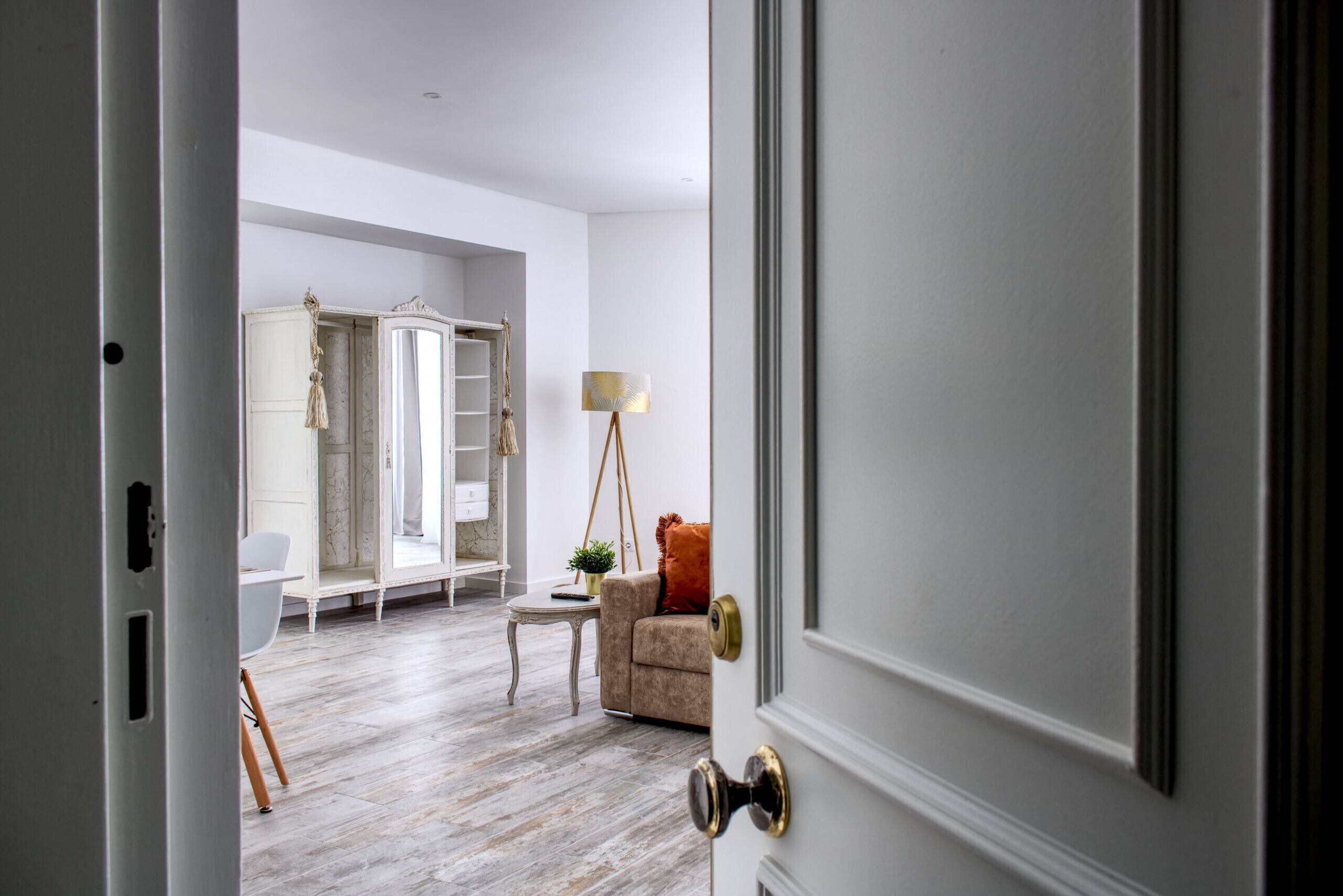 A room with a wooden floor, a white wardrobe, a beige sofa, a small table with a lamp, and a window with curtains.