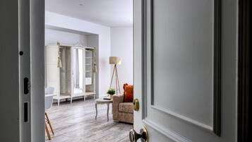 A room with a wooden floor, a white wardrobe, a beige sofa, a small table with a lamp, and a window with curtains.