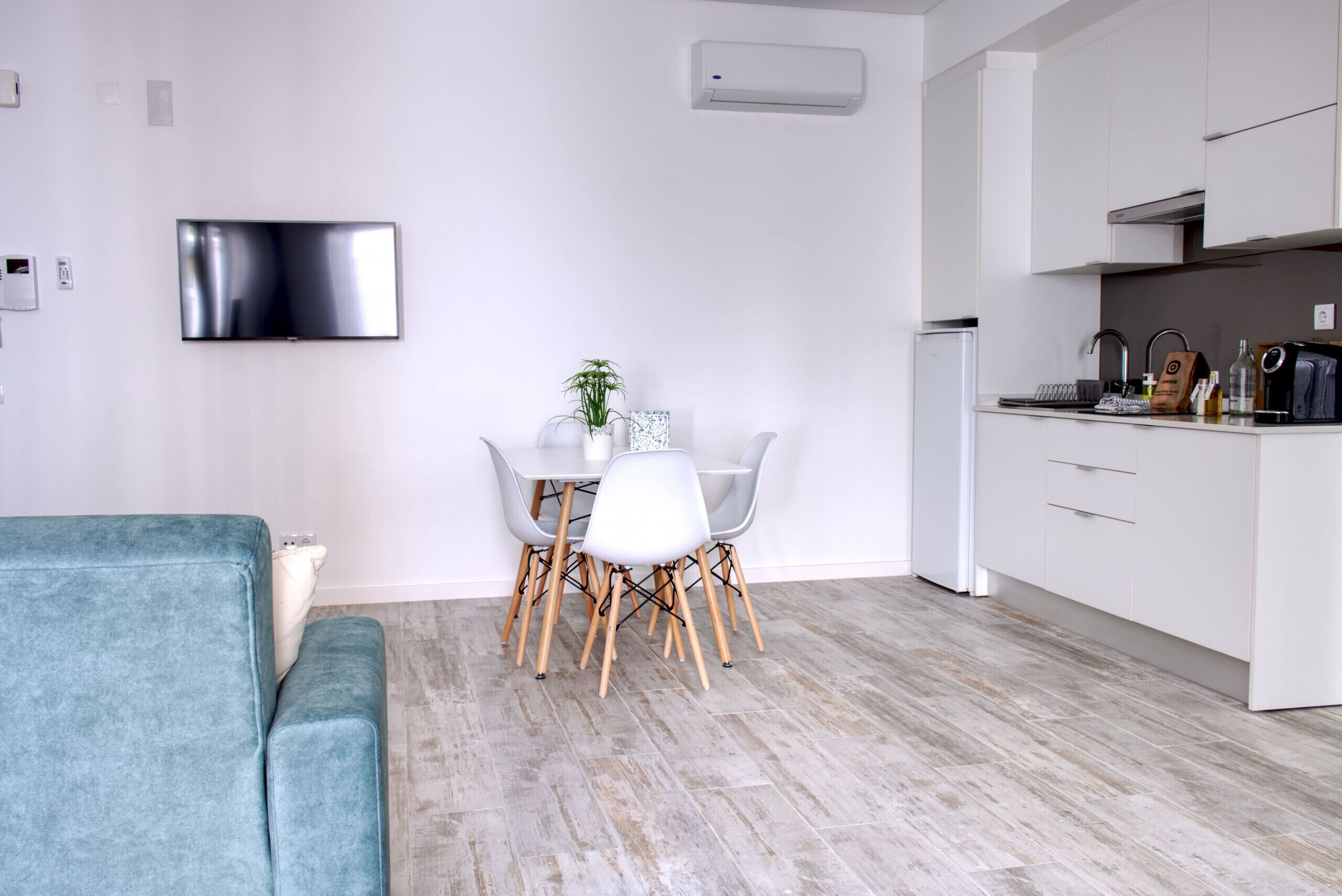 Un espace de vie moderne comprenant un canapé de couleur turquoise, une table à manger avec des chaises, un téléviseur à écran plat et un coin cuisine doté de meubles blancs et d’un four encastré.