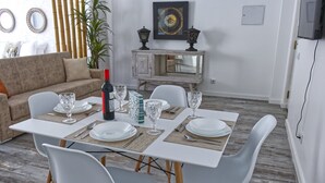 En matplats med ett vitt bord dukat för fyra personer, omgivet av vita stolar. I bakgrunden finns en soffa och ett skåp med dekorativa föremål.