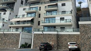 A multi-story residential building with balconies, a parking area, and a stone wall.