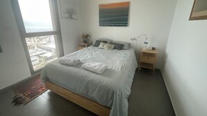 A bedroom with a large bed, a nightstand, a lamp, a painting, and a view of the city through a window.