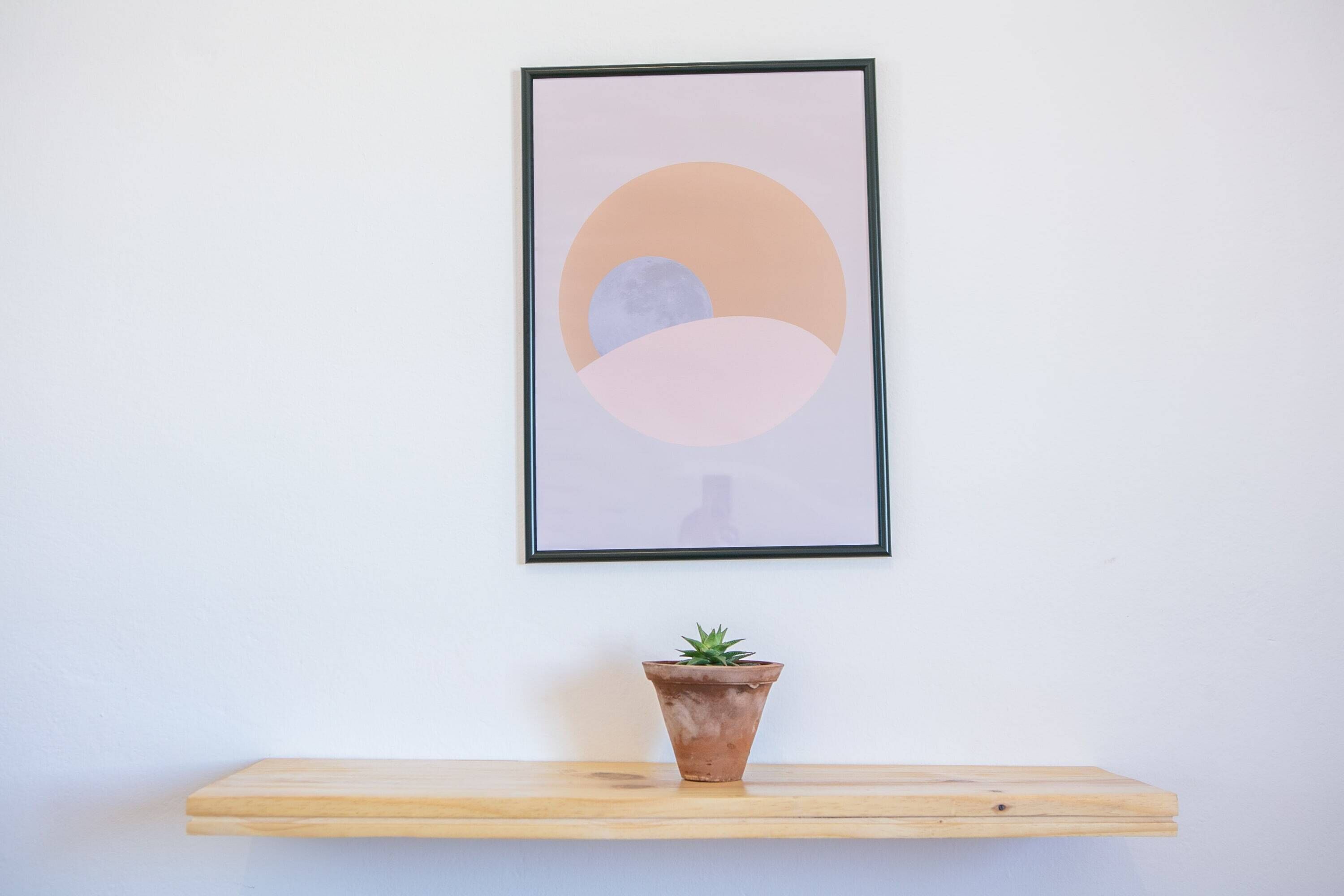 Ein gerahmtes abstraktes Kunstwerk an einer Wand über einem Holzregal mit einem Terrakottatopf und einer Pflanze.