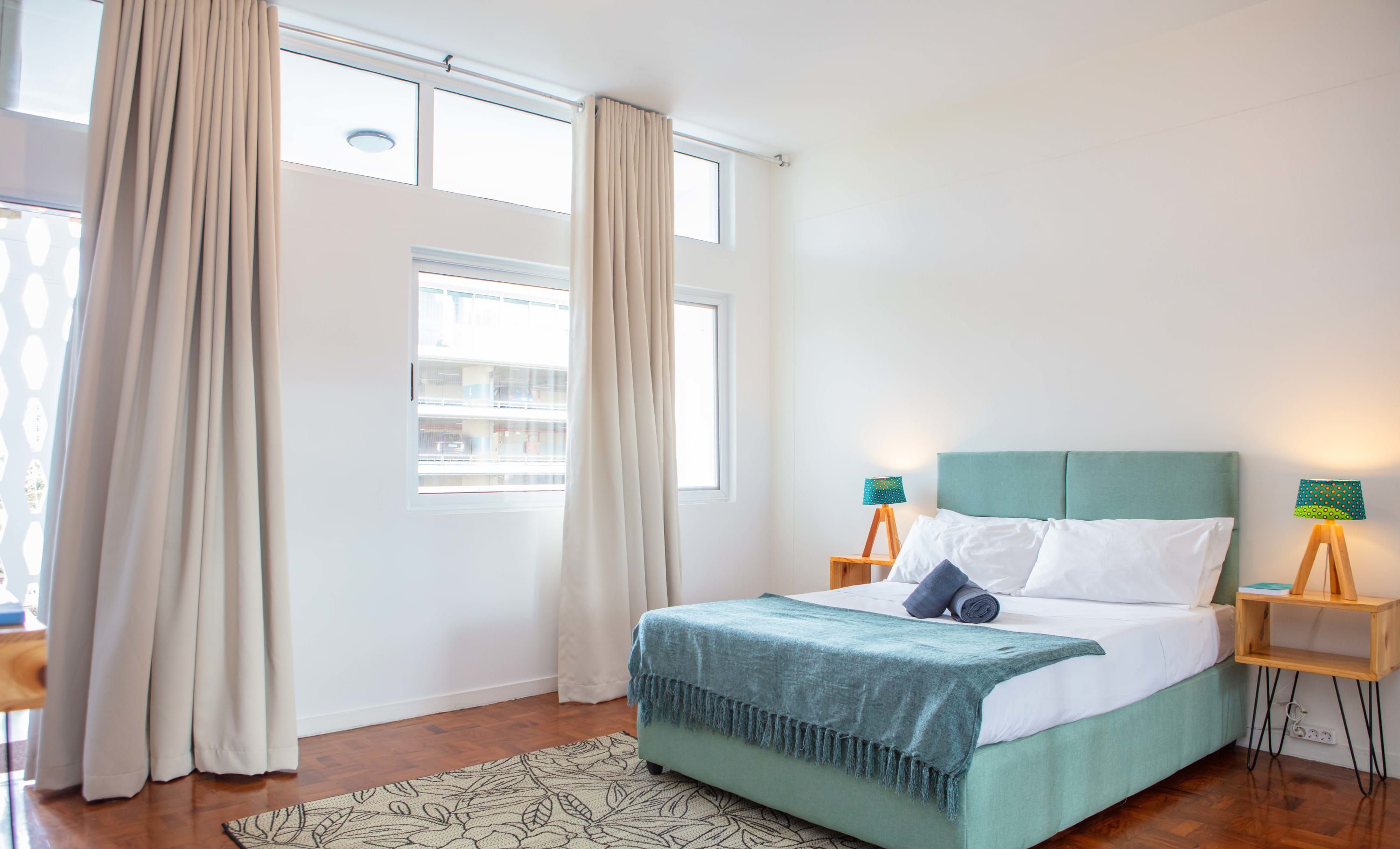 A bedroom with a bed, two bedside tables with lamps, a window with curtains, and a patterned rug.